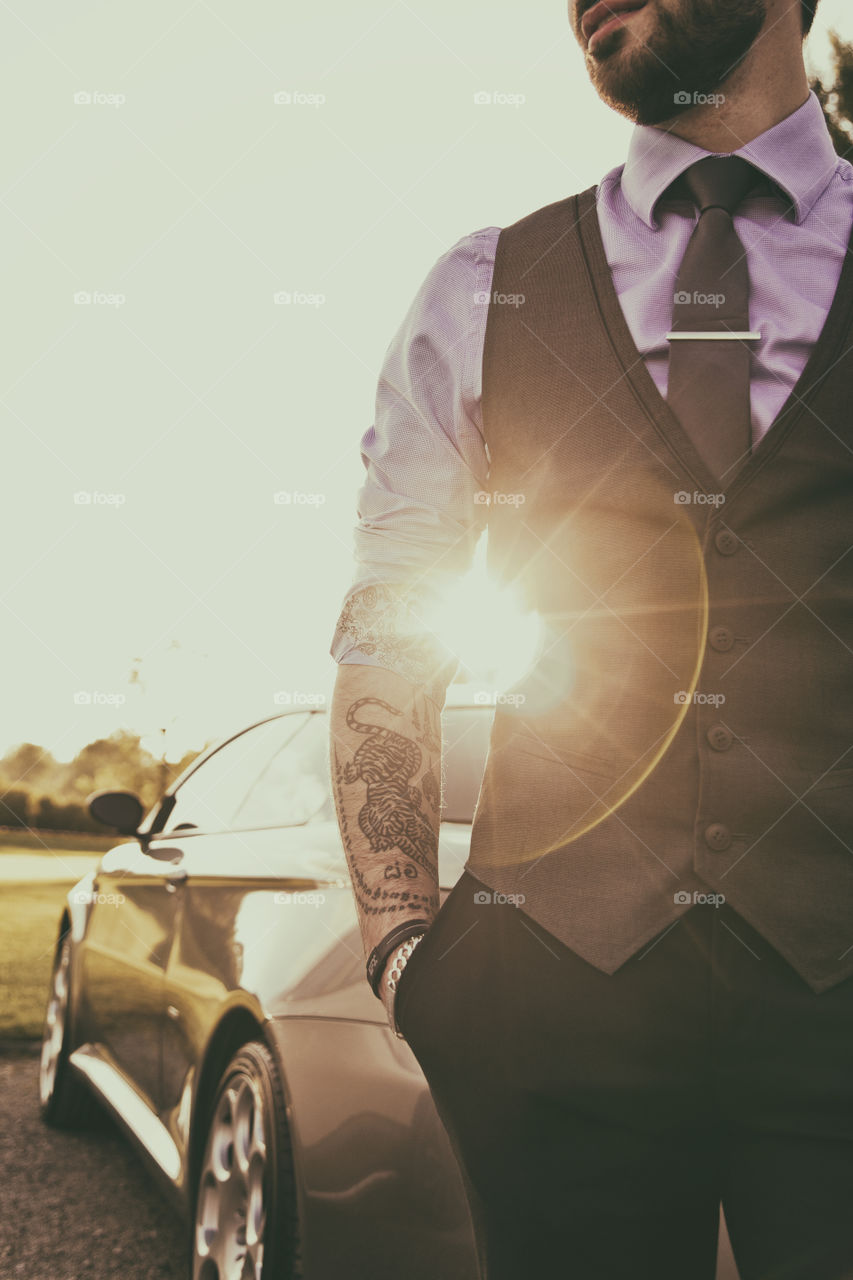 Portrait of a young man with tattoo of tiger and Thai words on arm wearing smart casual business clothing standing beside a sport car and sunrays
