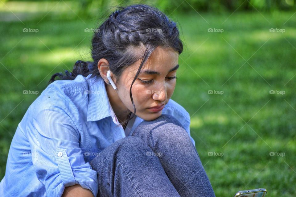 a girl in jeans and a shirt sits on a lawn in a park with a phone in her hands