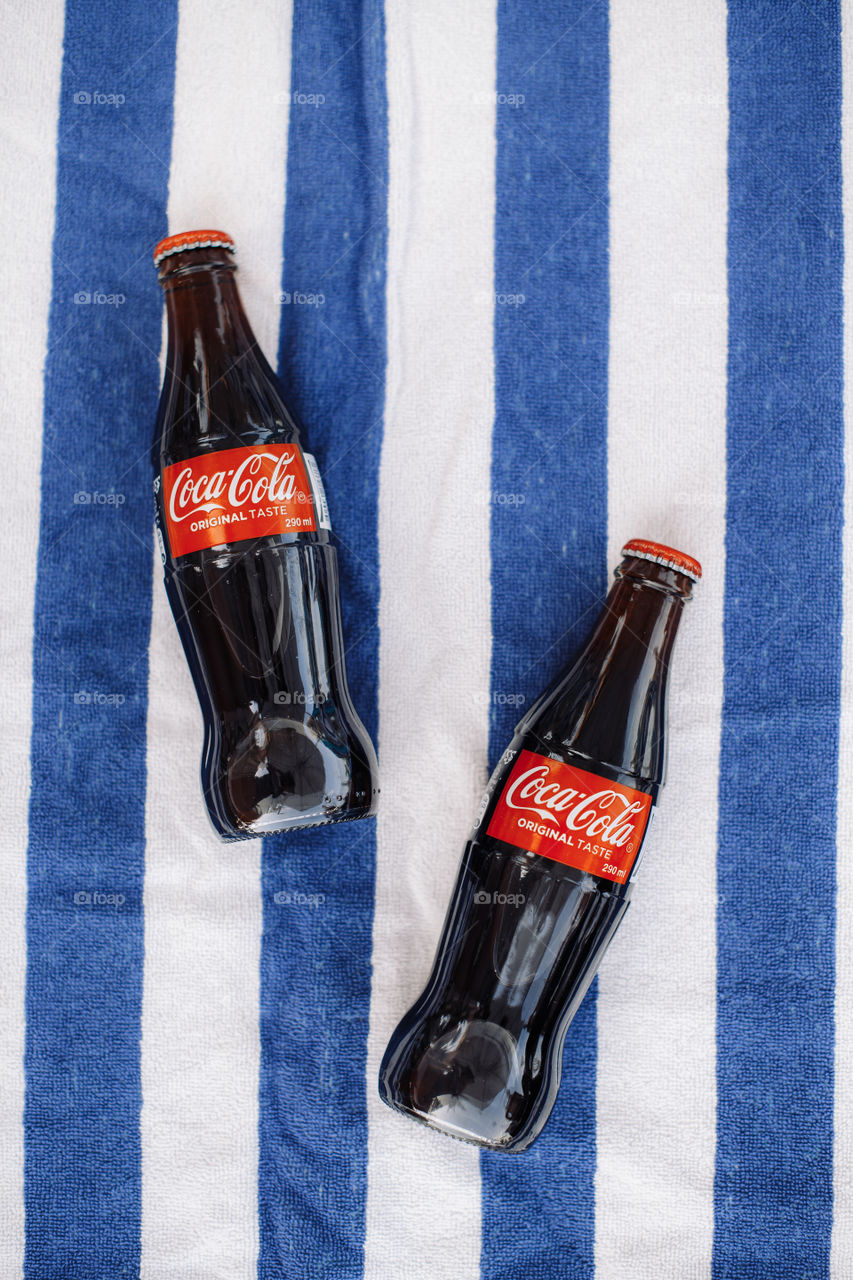 Two bottles of Coca-Cola lay on white blue towel