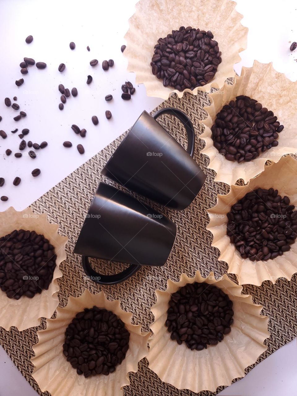 Coffee beans displayed in coffee filters surrounding two black coffee cups on beige placemat on a white surface.