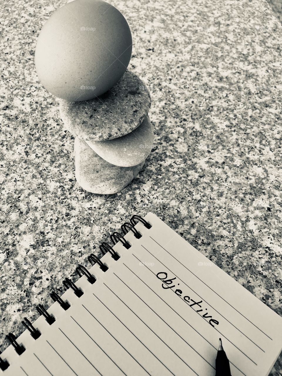 Whole intact hen’s chicken egg balancing atop smooth stone rock stack on marble surface, with spiral notebook and objective handwritten title, concept business, motivational, or life applications, black and white with copy space
