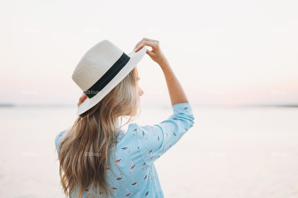 Beautiful blonde young woman in blue dress and straw hat on pier on the sunset