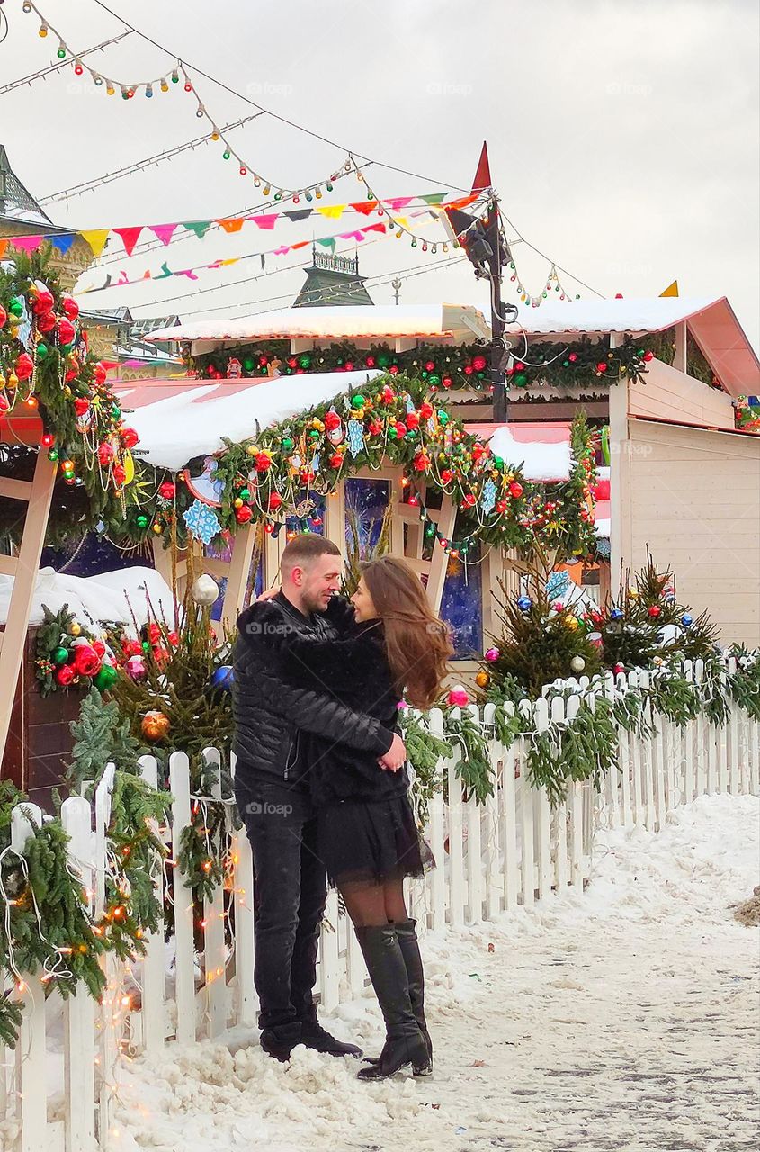 The main motivation of a person. Love, create a family and have children. A couple of lovers embrace in a snowy square. The look of lovers. Garlanded tents in the background