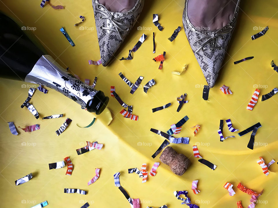 Female feet in fashionable shoes standing on yellow background with colorful confetti and opened bottle of champagne 