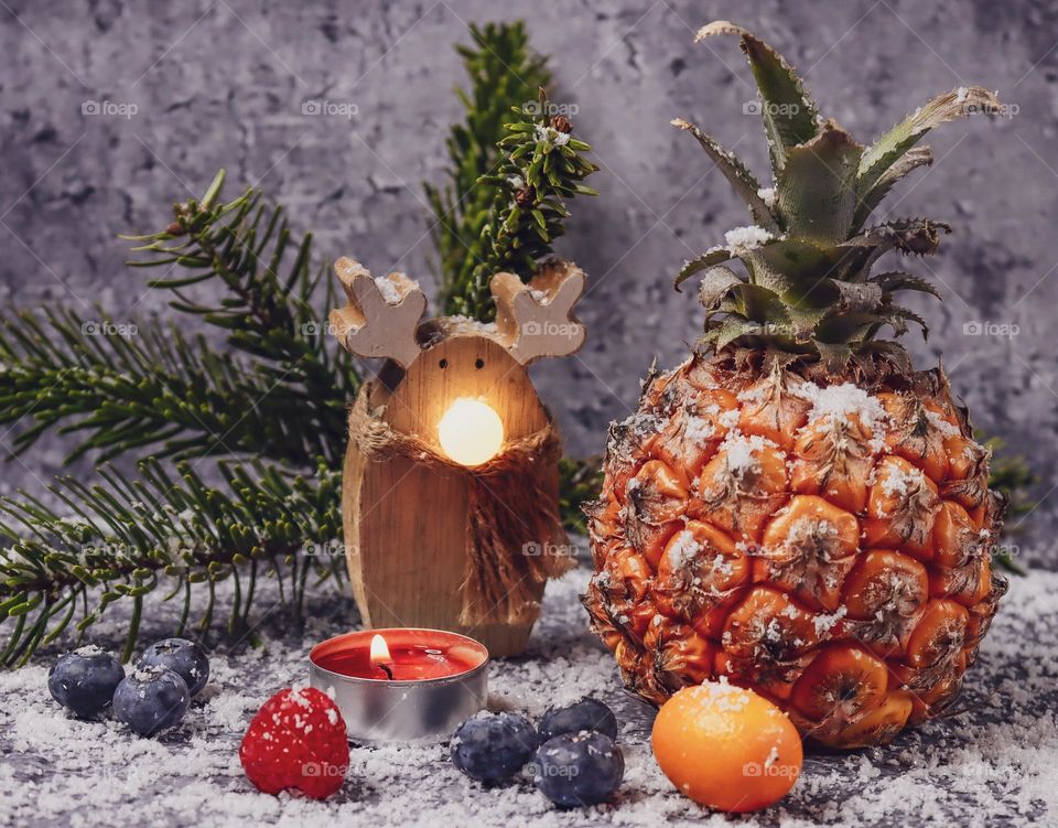 Tropical pineapple, raspberries, blueberries and mini tangerines with a Christmas tree branch and a wooden snowman with a luminous nose and sprinkled with snow on a dark cement background, close-up side view. The concept of Holiday fruits and berries