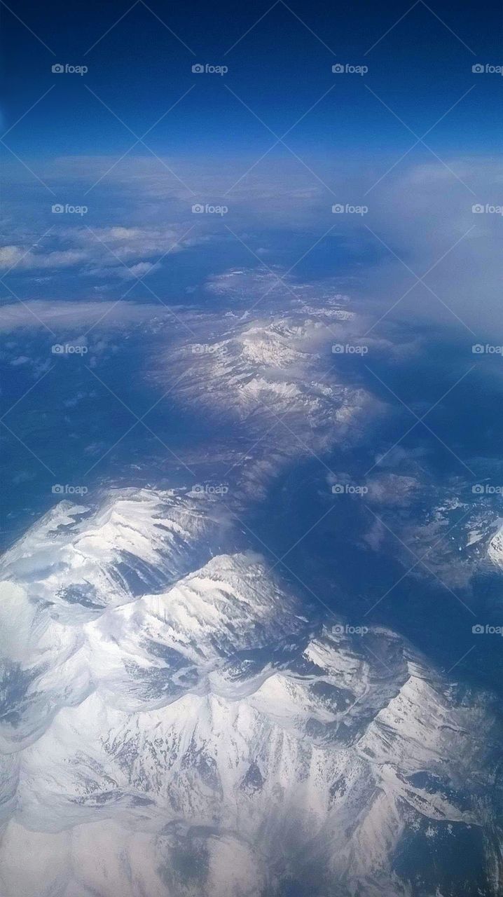 Aerial view of the snow-capped Rocky Mountains under a bright blue sky with a few clouds in between.