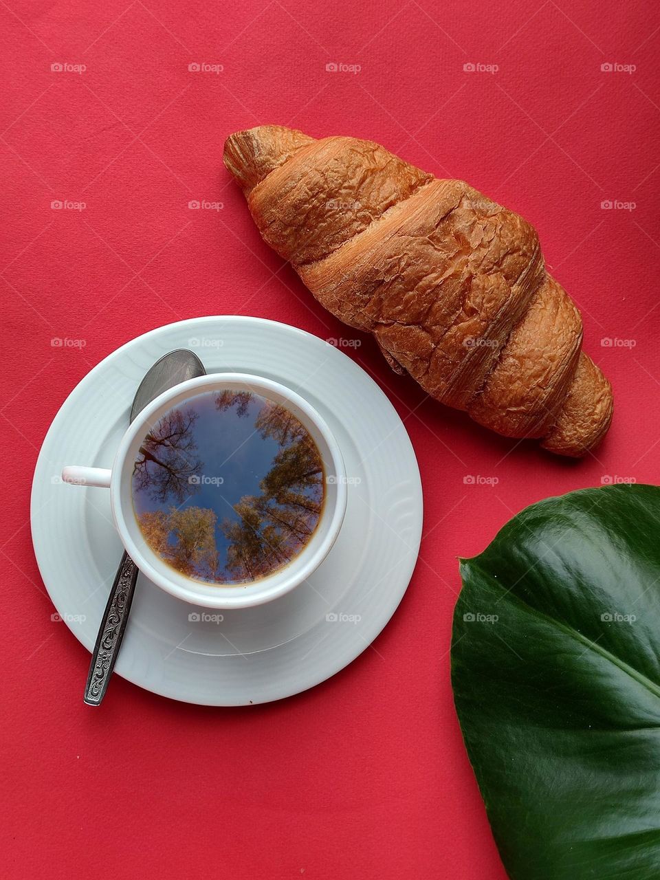 On a red background is a white cup on a white saucer with a metal spoon.  In the corner is a part of the green leaf of the Monstera plant.  A crispy croissant lies next to the cup.  The tops of the trees are reflected on the surface of the coffee