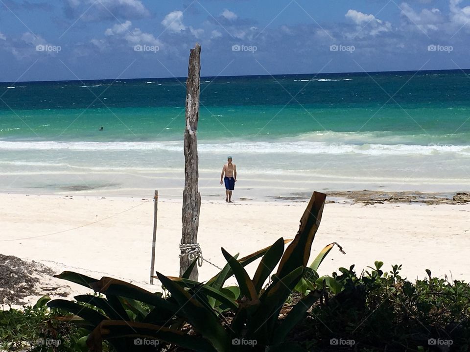 A man walks contemplatively on a white sand beach in Tulum, Mexico. Behind him, the crystal clear blue waters lap up at the shore. 