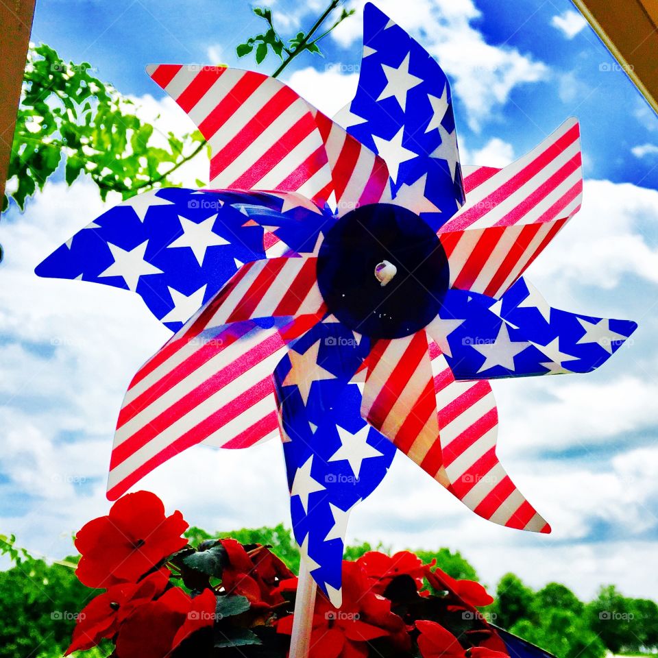 Patriotic Pinwheel against a blue sky and surrounded by red flowers. 